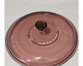 May include: A circular, translucent mauve glass lid featuring a smooth, flat surface with a raised, circular center section decorated with a radial, sunburst-like pattern and a small, dark purple knob handle at the very top center.