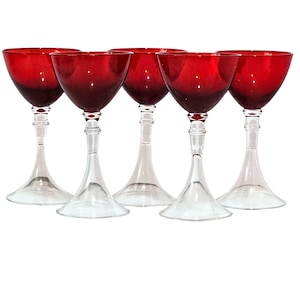 May include: Five stemmed wine glasses with ruby red bowls and clear stems. The glasses have a classic design, with a wide bowl that tapers to a narrow stem and a flared base. The glasses are arranged in a row against a white background.