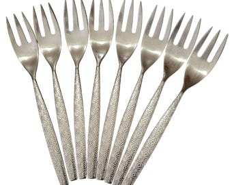 May include: A set of eight silver-toned metal cocktail forks arranged in a fan shape. Each fork features three tines and a slender handle decorated with a repeating embossed geometric pattern of small circles and intersecting lines along the stem.