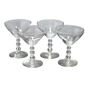 May include: A set of four clear glass coupe glasses, each featuring a wide, shallow bowl and a stem composed of three stacked spherical glass beads, resting on a circular base. The glassware is transparent and designed for serving cocktails.