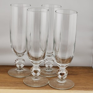 May include: Four clear champagne flutes with heart-shaped bases. The glasses have a tall, slender design, perfect for celebratory occasions. The heart-shaped detail adds a touch of elegance to the stemware. The flutes are displayed on a wooden surface.