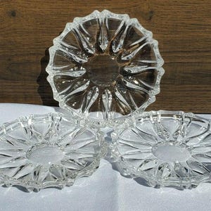 May include: Three clear glass ashtrays with a starburst design. The ashtrays have a circular shape with a textured, ridged rim. The ashtrays are made of clear glass and are displayed on a white surface with a wooden background.