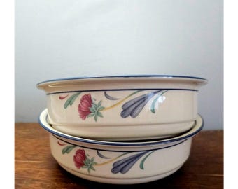May include: Two stacked off-white ceramic bowls featuring a dark blue rim and a painted floral pattern. The design includes stylized pink flowers with green leaves and elongated blue petals arranged in a repeating motif around the exterior.