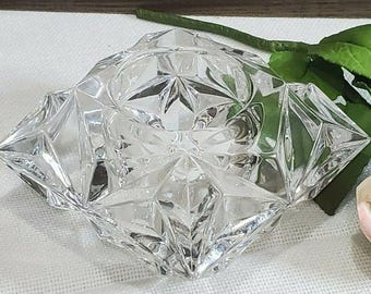 May include: A clear, faceted glass candle holder with a square base and a circular center opening. The exterior surface features a geometric, multi-angled design that reflects light, creating a prismatic effect across the transparent glass structure.