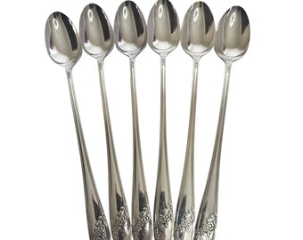 May include: A set of six long-handled silver-toned spoons arranged in a vertical row. Each spoon features a polished oval bowl and a slender handle decorated with a detailed floral relief pattern at the base, showcasing a reflective metallic finish.