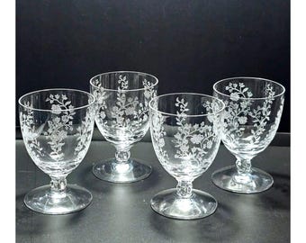 May include: A set of four clear glass goblets, each featuring a delicate etched floral pattern with vines and blossoms. The glasses have a rounded bowl, a short stem with a small decorative knob, and a wide, flat circular base for stability.
