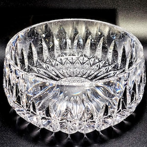 May include: A clear, round crystal bowl with intricate, geometric patterns. The bowl's design features a series of pointed shapes and radial lines, creating a textured surface. The bowl is likely used for serving or display.