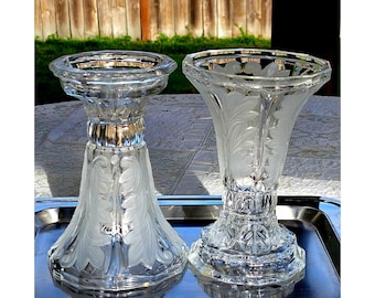 May include: Two clear glass candle holders featuring a frosted leaf pattern etched into the surface. Each holder has a wide, flared top rim and a sturdy, multi-faceted base. The glass is transparent with matte white frosted detailing.