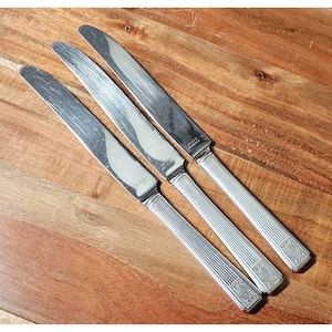 May include: Four stainless steel knives with long, slender blades and ridged handles. The knives are arranged diagonally on a wooden surface. The handles have a decorative diamond pattern at the end.