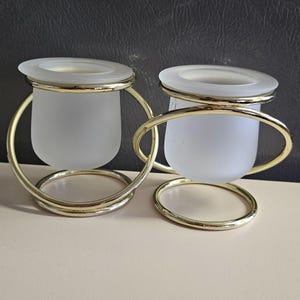 May include: Two frosted glass candle holders with gold metal rings. The candle holders are on a light beige surface.