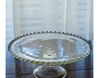 May include: A clear glass cake stand featuring a wide, flat circular plate with a decorative beaded edge. The stand is supported by a sturdy, fluted pedestal base that flares outward at the bottom, providing a stable foundation for the display.