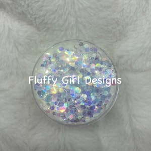 Pode incluir: Um recipiente transparente cheio de glitter iridescente em tons de branco, azul e roxo. O recipiente tem o texto "Fluffy Girl Designs" impresso no lado.