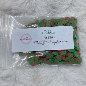 Puede incluir: Una bolsa de plástico transparente llena de espolvoreos en forma de duende verde y rojo. La bolsa tiene una etiqueta blanca que dice "Goblin (No comestible) ThatGlitterSupplier.com".