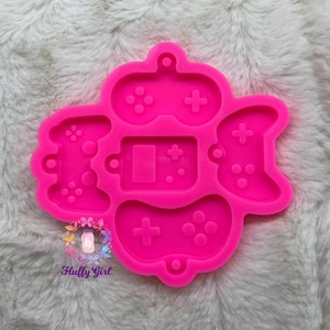 May include: Pink silicone mold with four different video game controller designs. The mold is shaped like a heart and has a small hole at the top for hanging.