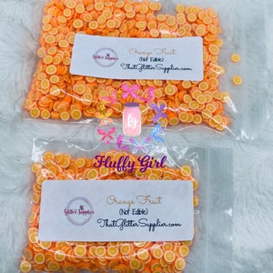 May include: Two clear plastic bags filled with orange fruit slices. Each slice is orange with a yellow center. The bags have white labels with the text "Orange Fruit (Not Edible) ThatGlitterSupplier.com". The brand name "Fluffy Girl" is also visible.