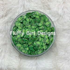 Puede incluir: Un cuenco de vidrio transparente lleno de abalorios de resina verde en forma de cara de extraterrestre. Los abalorios son planos y tienen ojos negros. El cuenco está sobre una superficie blanca y esponjosa. "Fluffy Girl Designs" está escrito en negro en el cuenco.