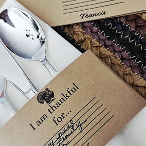 Puede incluir: Tarjetas de mesa con temática de Acción de Gracias con una ilustración de pavo y el texto "Estoy agradecido por...". Las tarjetas están sobre una servilleta blanca con cubiertos.