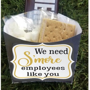 以下が含まれることがあります： 「We need S'more employees like you」と金色の文字で書かれた白黒の看板。看板は、グラハムクラッカー、チョコレートバー、マシュマロが入った黒い紙の円錐形の中にあります。