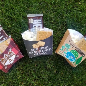 May include: Three brown paper bags filled with ingredients for making s'mores. The bags are decorated with different camping-themed designs and text. One bag says "Let's Get Toasted", another says "Where friends and marshmallows get toasted", and the third says "Happy Campers".