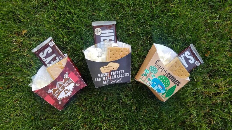 May include: Three brown paper bags filled with ingredients for making s'mores. The bags are decorated with different camping-themed designs and text. One bag says "Let's Get Toasted", another says "Where friends and marshmallows get toasted", and the third says "Happy Campers".