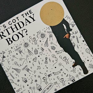 May include: A birthday card with the text "Who's Got The Birthday Boy?" and a scratch-off circle. The card features a black and white doodle design with party-themed illustrations, including cupcakes, balloons, and confetti.