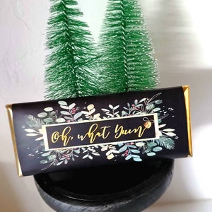 May include: A black and gold chocolate bar with a floral design and the text "Oh, what yum" on a black pedestal with two small green artificial Christmas trees in the background.
