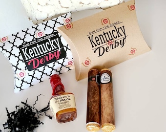 Puede incluir: Un set de regalo de Kentucky Derby con una caja de patrón blanco y marrón, una botella en miniatura de whisky Maker's Mark, dos cigarros marrones y una bolsa de papel marrón con el texto "RUN FOR THE ROSES Kentucky Derby" impreso en ella.