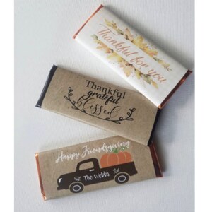 May include: Three chocolate bar wrappers with Thanksgiving themes. One wrapper has a fall leaf design with the text "Thankful for you". Another wrapper has a brown background with the text "Thankful grateful blessed". The third wrapper has a brown background with a black silhouette of a pickup truck carrying a pumpkin and the text "Happy Friendsgiving The Webbs".