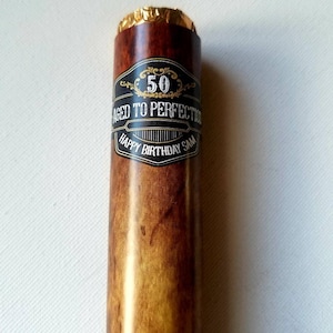 Puede incluir: Un tubo de cigarro marrón y dorado con una etiqueta negra que dice "50 Aged to Perfection Happy Birthday Sam".