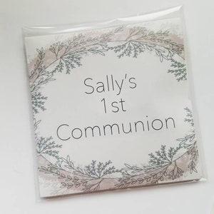Puede incluir: Una tarjeta cuadrada dentro de una funda de plástico transparente con el texto Sally's 1st Communion en letras negras. La tarjeta está decorada con una delicada ilustración de corona botánica circular en tonos gris suave y rosa.
