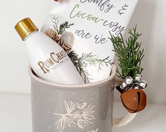May include: A gray ceramic mug with the text "Comfy & Cozy" printed on it. The mug contains a small bottle of rumchata liqueur, a bag of cocoa mix with the text "Comfy & Cocoa-cozy are we" printed on it, a cinnamon stick, and a small sprig of greenery with a silver bell.