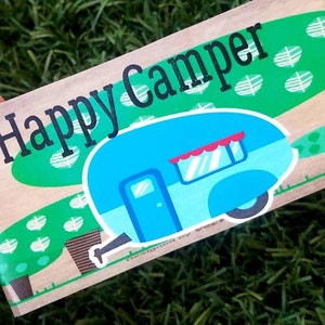May include: A brown wooden sign with the text "Happy Camper" in black lettering. The sign features a cartoon illustration of a blue camper van with a red awning and white wheels, set against a green background with white leaf designs.