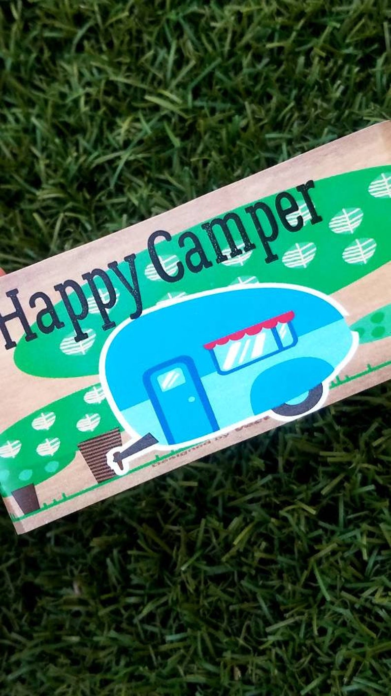 May include: A brown wooden sign with the text "Happy Camper" in black lettering. The sign features a cartoon illustration of a blue camper van with a red awning and white wheels, set against a green background with white leaf designs.