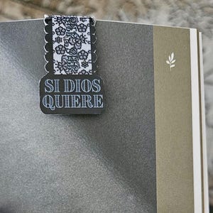 May include: A black and white bookmark with the text "SI DIOS QUIERE" in white lettering. The bookmark has a floral patterned ribbon and is clipped onto the pages of a book.