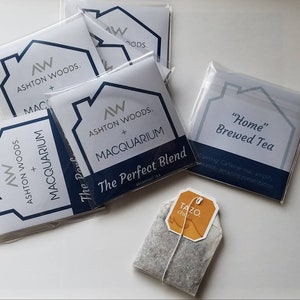May include: A set of five white square cards with blue and silver text. The cards feature a house-shaped logo with the text "Ashton Woods + Macquarium" and "The Perfect Blend". One card has the text "Home Brewed Tea" with the text "Warming Caffeine, a jolt of fresh amazing presentation". A single tea bag with a yellow label that says "TAZO chai" is in the lower right corner of the image.