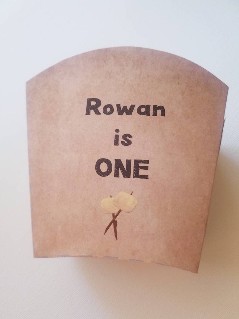 May include: A brown paper party favor bag with the text "Rowan is ONE" and a graphic of two marshmallows on sticks.