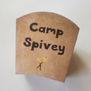 May include: A brown paper cup with the text "Camp Spivey" printed in black. There are two yellow marshmallow drawings on sticks.