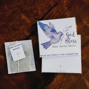 May include: A white tea bag with a blue butterfly pea flower tea tag. The tag features a watercolor illustration of a dove with an olive branch in its beak. The text on the tag reads "God Bless Baby Xavier Becket" and "Blue Butterfly Pea Flower Tea". The date "12.29.22" is printed on the bottom of the tag.