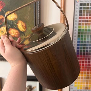 May include: A cylindrical ice bucket featuring a dark brown wood grain exterior, a polished silver-tone metal lid, and a matching silver-tone metal swing handle. A small, circular, dark brown wooden knob is centered on top of the lid.