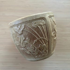 May include: A beige ceramic planter with a textured surface and embossed floral design. The planter has a square shape with rounded corners and a slightly flared rim. The floral pattern includes leaves and stems.