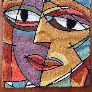 May include: A square textile wall hanging featuring a multi-colored, abstract face design. The piece is composed of various geometric shapes in shades of purple, gold, red, blue, and green, all outlined in thick black thread with a textured finish.