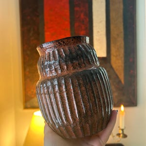 May include: A brown and black ceramic vase with vertical ribbed detailing. The vase has a rounded shape with a slightly flared top. The color gradient transitions from brown to black. The vase is held in a hand.