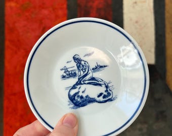 May include: Small white plate with a blue rim and a blue illustration of a mermaid on a rock. The plate has a classic design, suitable for serving small portions or as a decorative piece.