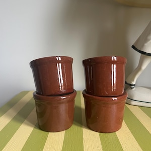 May include: Four small, brown ceramic ramekins stacked on a green and yellow striped surface. The ramekins have a simple, rounded design with a slightly flared rim. The glossy finish reflects light.