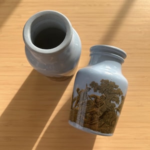 May include: Two light blue ceramic vases. One vase has a detailed gold-colored illustration of a hunting scene. The vases have a cylindrical shape with a slightly flared rim. The background is a light brown wooden surface.