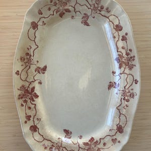 May include: An oval-shaped, antique serving platter with a cream-colored base. The rim is decorated with a repeating pattern of burgundy-colored floral vines and berries. The platter has a slightly irregular, scalloped edge.