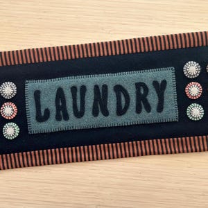 May include: A rectangular laundry sign with a black background and a brown and orange striped border. The word "LAUNDRY" is stitched on a gray rectangle in the center. Decorative buttons in white, pink, and green are arranged around the sign.