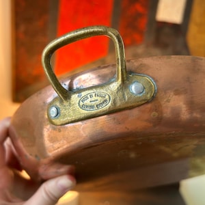 May include: A copper-colored pan with a brass handle and rivets. The pan has a rounded shape and a shiny surface. The brass handle is attached to the pan with rivets. The text "MADE IN FRANCE" is visible on the pan.