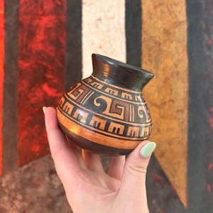 May include: A small, rounded ceramic pot featuring a dark black rim and base with a central band of orange clay. The surface is decorated with intricate, hand-painted geometric patterns in black, including repeating step and spiral motifs.