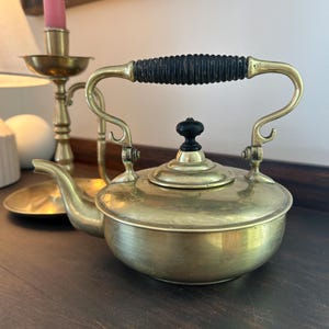 May include: Vintage brass teapot with a black handle and lid finial. The teapot has a curved spout and a hinged handle. A brass candlestick with a pink candle is in the background.